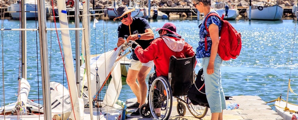Personne en fauteuil sur un ponton en train de gréer son miniji avec l'aide de 2 bénévoles